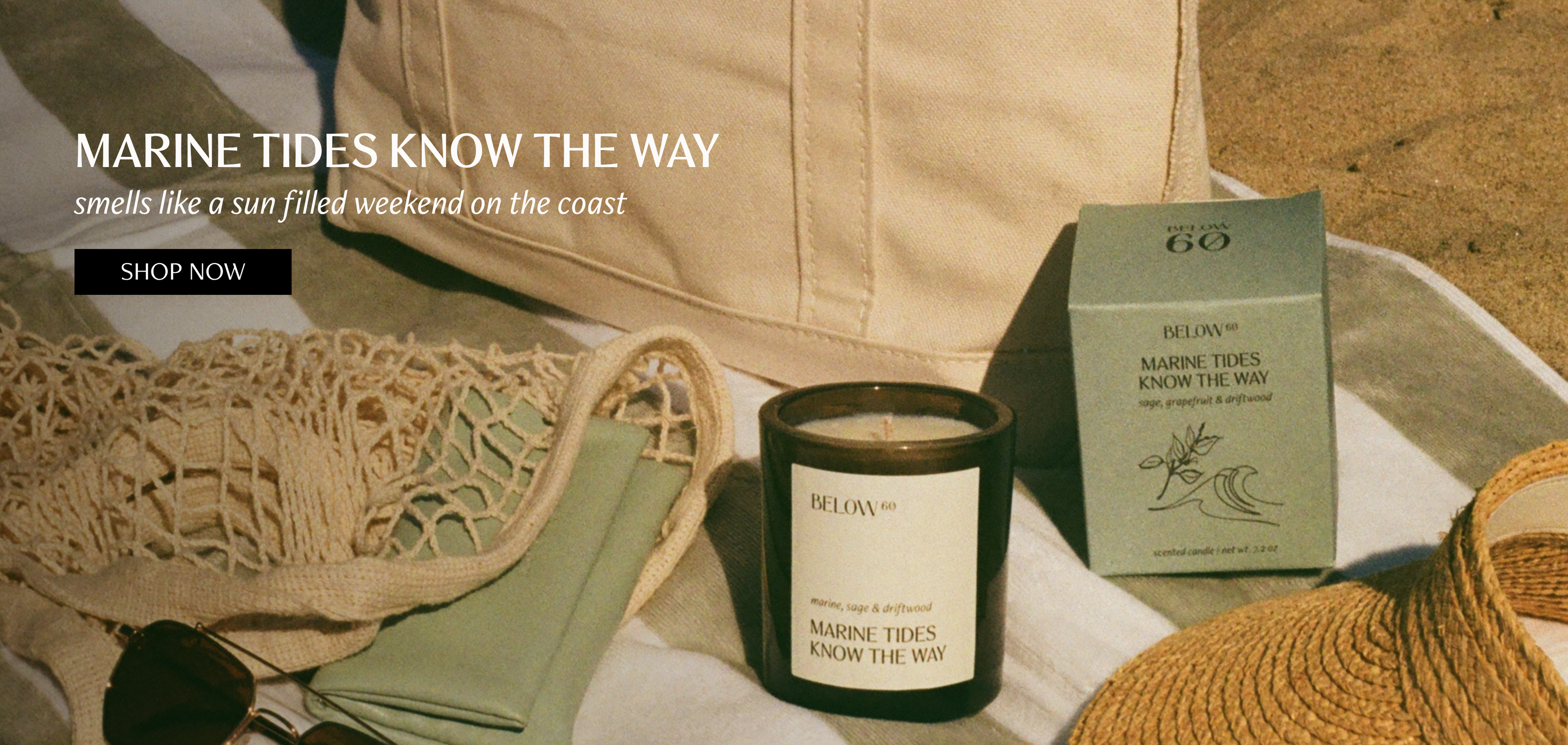 A canvas tote bag and woven straw accessories on a striped beach towel with a Below 60 Marine Tides Know the Way candle and its packaging box, styled in warm, sun-faded tones.