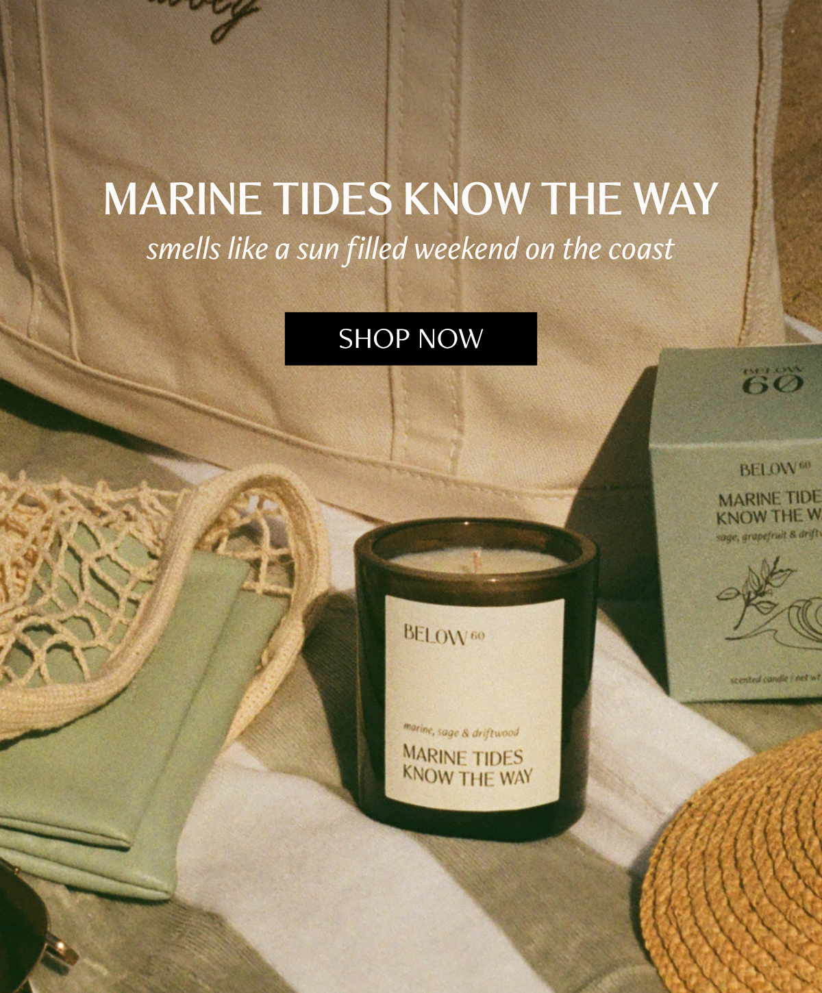 A Below 60 Marine Tides Know the Way candle in a dark glass vessel sits on a striped beach towel beside a canvas tote, mesh beach bag, and the candle's packaging box, with straw and sunglasses visible at the edges.