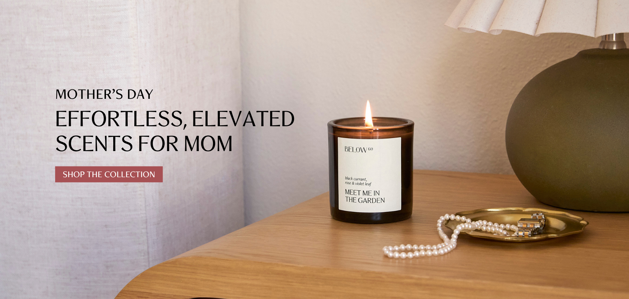 A lit Below 60 Meet Me in the Garden candle in a dark amber jar sits on a warm wood surface beside a pearl necklace and gold jewelry dish, with a linen curtain and sculptural lamp in the background. Overlay text reads Mother's Day — Effortless, Elevated Scents for Mom.