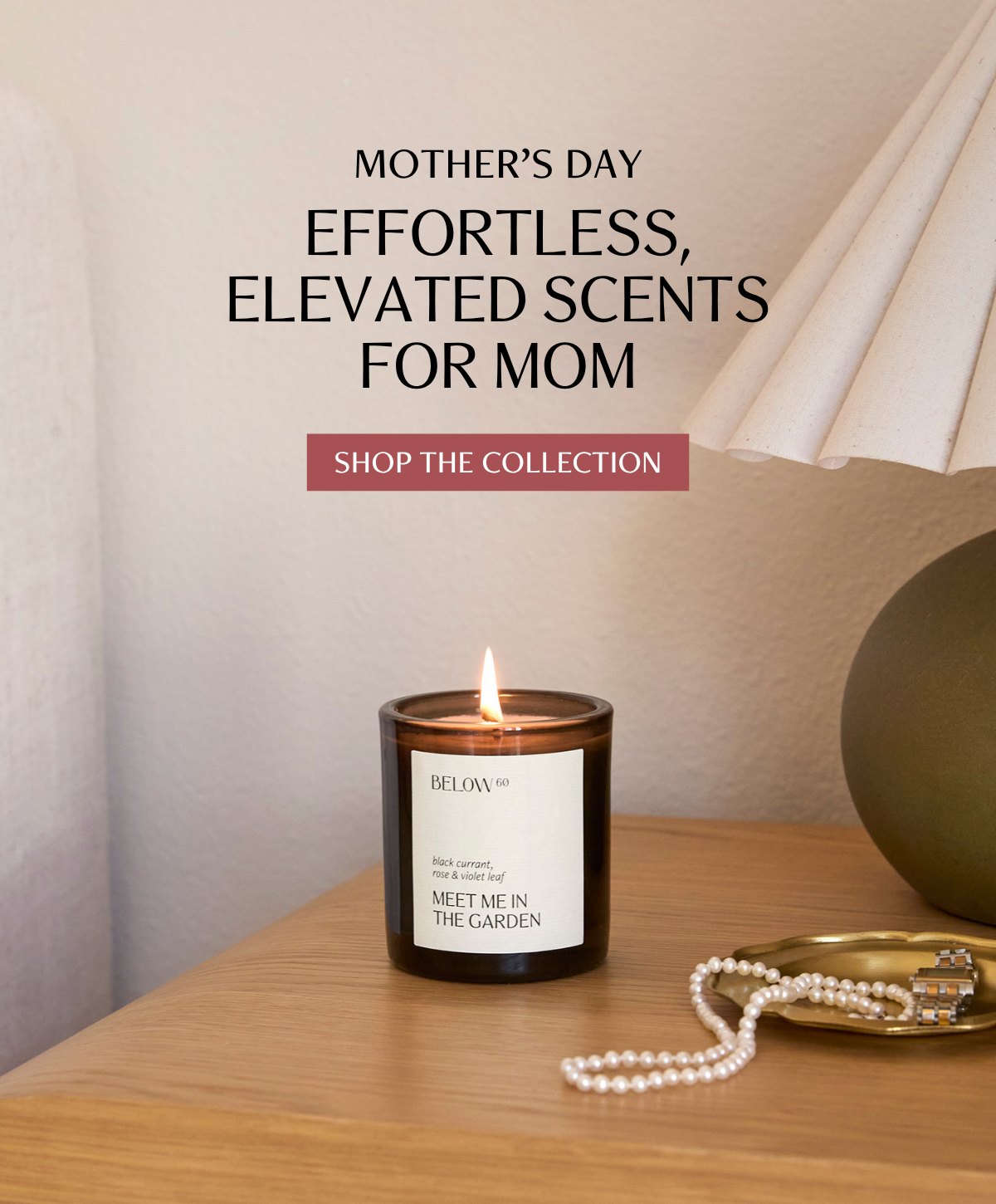 A lit Below 60 Meet Me in the Garden candle in a dark amber jar sits on a warm wood surface beside a pearl necklace and gold jewelry dish, with a sculptural lamp in the background. Overlay text reads Mother's Day — Effortless, Elevated Scents for Mom.