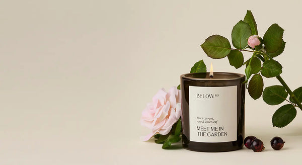 Candle with a label 'Meet Me in the Garden' surrounded by flowers and leaves on a beige background
