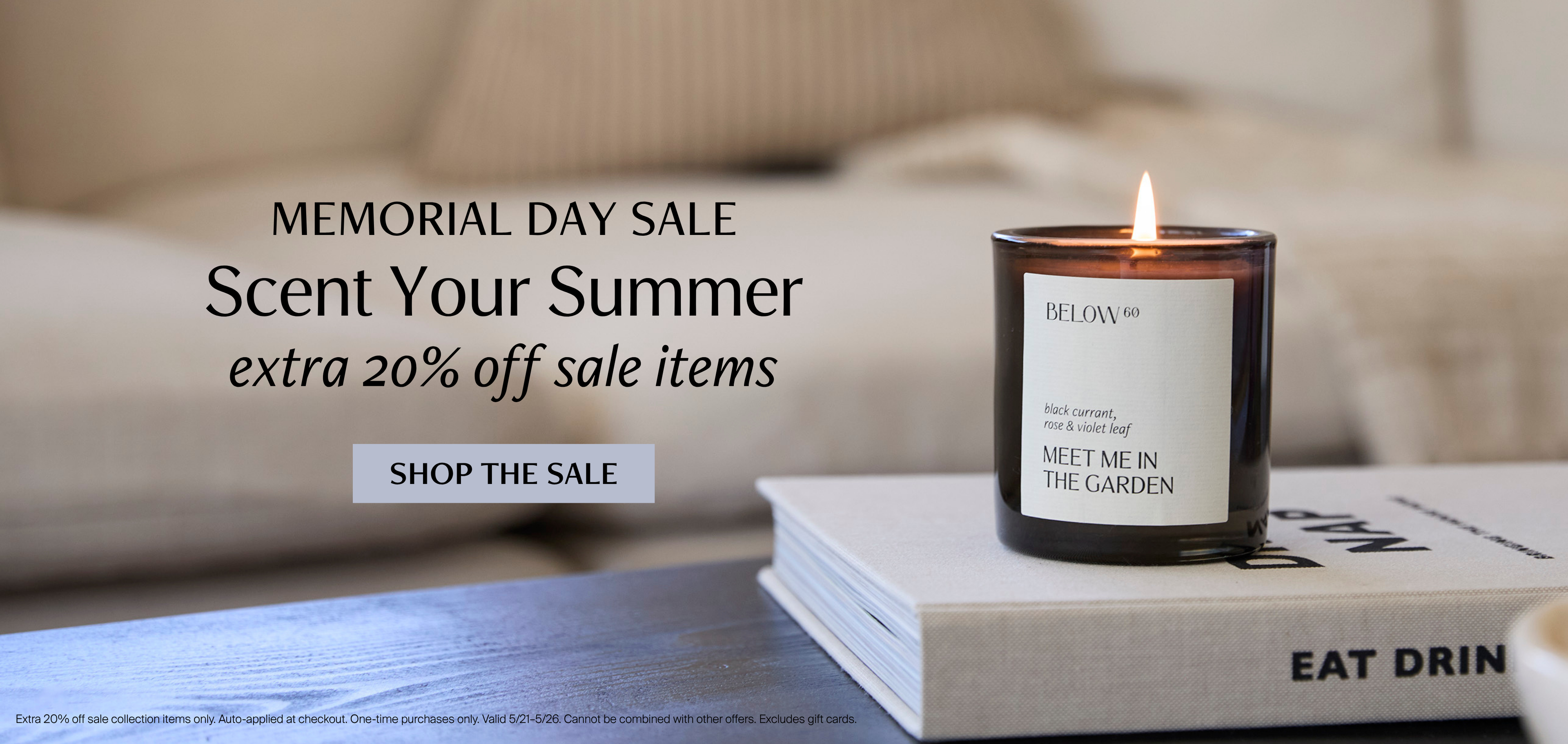 A lit Meet Me in the Garden candle by Below 60 resting on a stack of coffee table books against a soft neutral linen background. Text reads: Memorial Day Sale — Scent Your Summer, extra 20% off sale items.